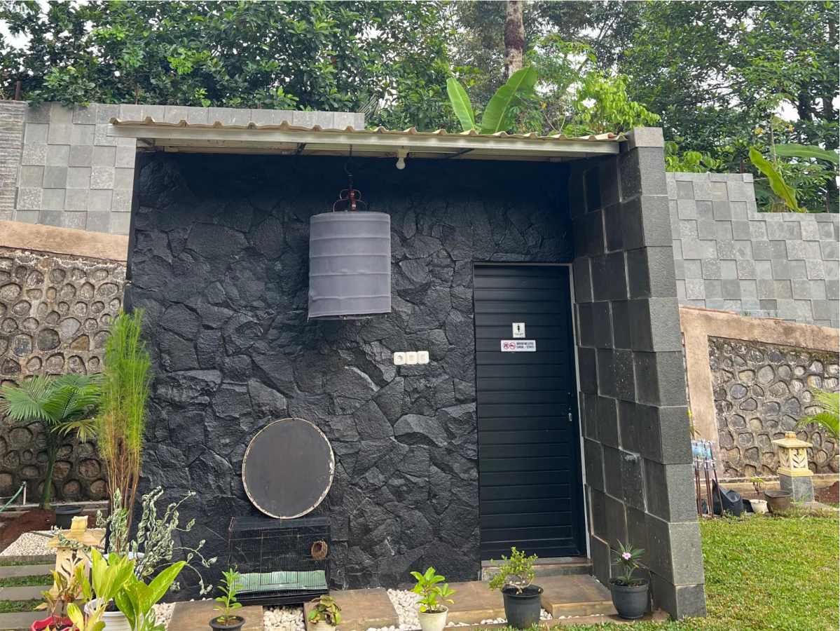 Fasad bangunan WC outdoor berwarna gelap dengan aplikasi batu templek hitam acak pada dinding utama dan batu andesit bakar pada pilar penyangga.