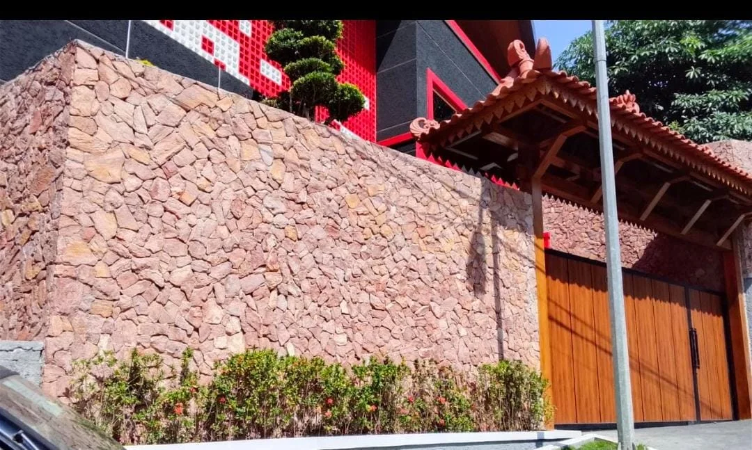 Pagar dinding luar berukuran besar yang dilapisi penuh oleh batu alam sandstone merah dengan potongan acak, bersanding dengan pintu gerbang aksen kayu dan area penghijauan di bagian bawah.