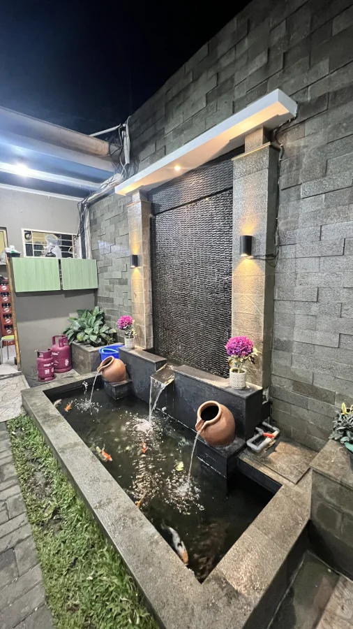 Kolam ikan koi bergaya minimalis dengan dinding berlapis batu andesit bakar abu-abu, dilengkapi ornamen air terjun dinding, pancuran kendi tanah liat, dan lampu hias dinding.